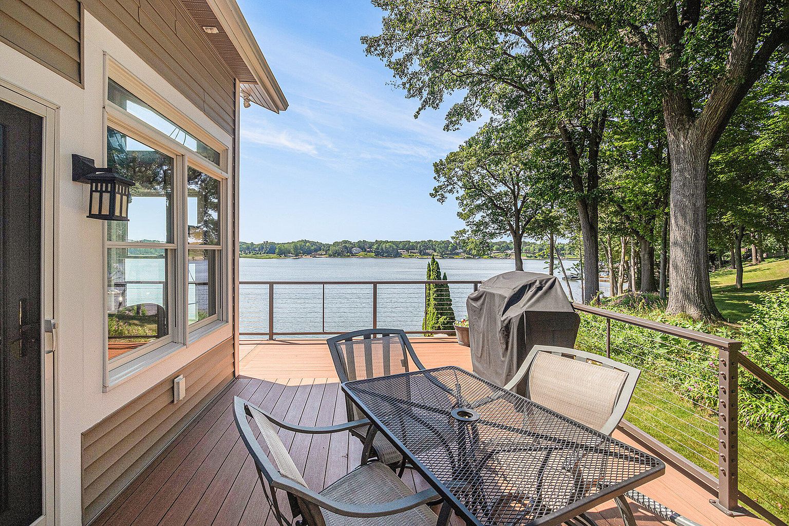 This image showcases a beautiful outdoor deck area with a view of a serene lake. The deck is furnished with a metal table and chairs, and a grill is positioned nearby, suggesting an ideal space for outdoor dining and relaxation. The deck features a modern railing system, and the surrounding trees add a touch of nature and privacy.