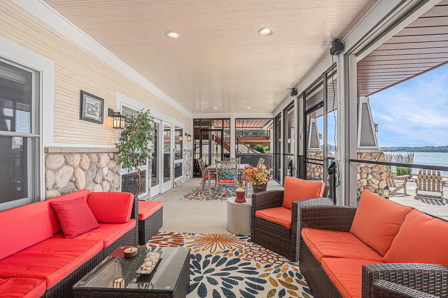 This is a beautifully furnished outdoor patio or sunroom, featuring comfortable wicker seating with vibrant red and orange cushions. The space is decorated with colorful rugs and a glass-topped coffee table, creating a cozy and inviting atmosphere. Large windows offer scenic views of the water, enhancing the appeal of this outdoor living area.