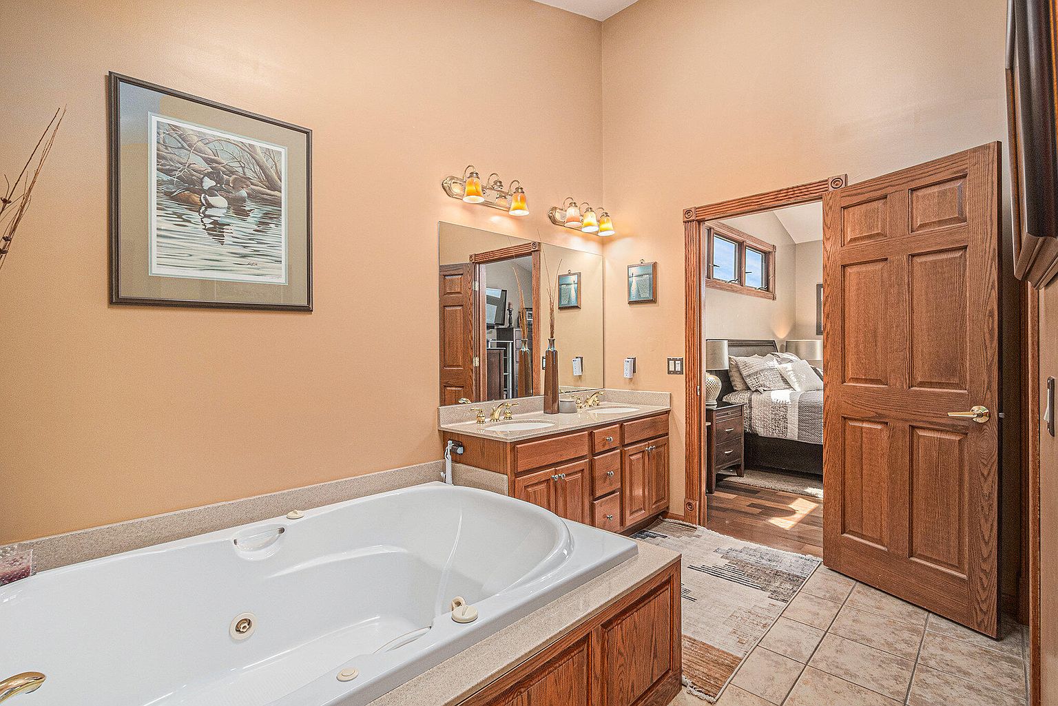 This is a primary bathroom featuring a large jacuzzi tub with wood paneling, a double vanity with wood cabinets and granite countertops, and a doorway leading to the primary bedroom. The walls are painted in a warm, neutral tone, and the flooring is tiled. The overall impression is luxurious and inviting.