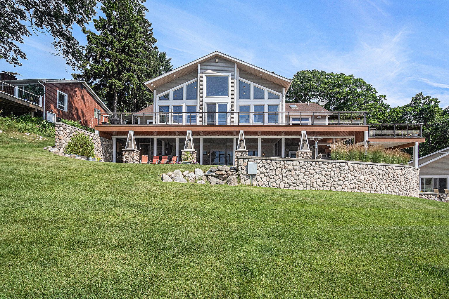 This is a rear view of a modern two-story house with a large deck. The house features a wall of windows on the upper level and a stone retaining wall in the yard. The lawn is well-maintained and lush, creating an inviting outdoor space.