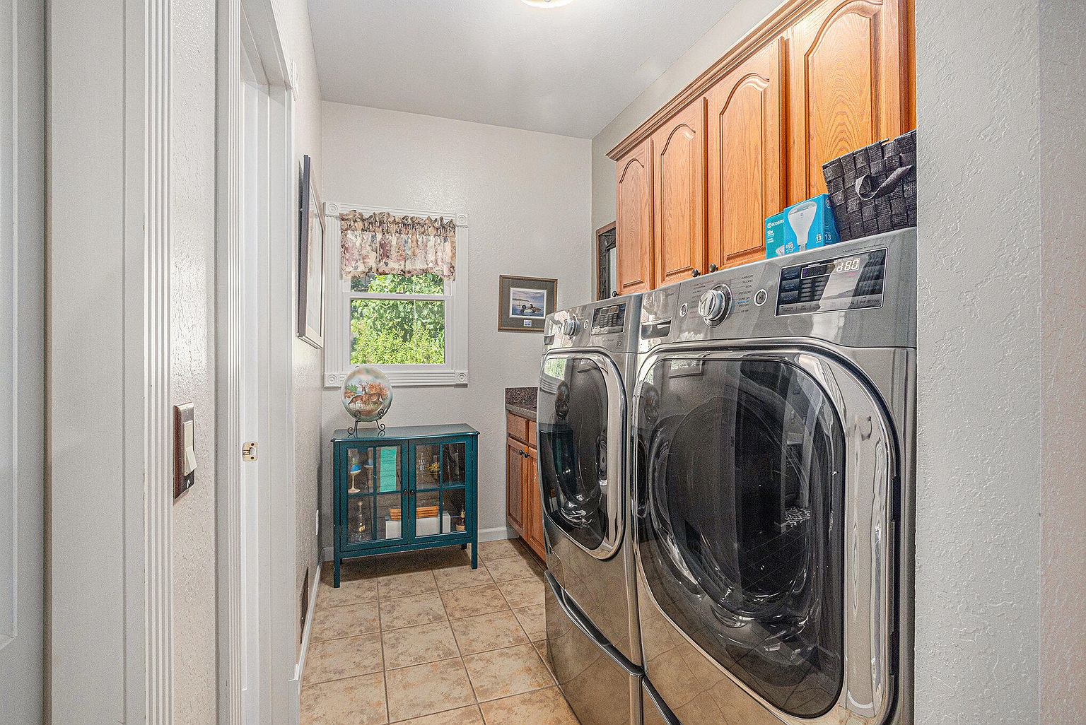 This is a well-organized laundry room featuring a stacked washer and dryer set in a sleek, modern design. Wooden cabinets above the appliances provide ample storage space. A small window with a decorative valance allows natural light to filter into the room, and a teal cabinet adds a pop of color. The perspective is from the doorway, showcasing the depth of the room.