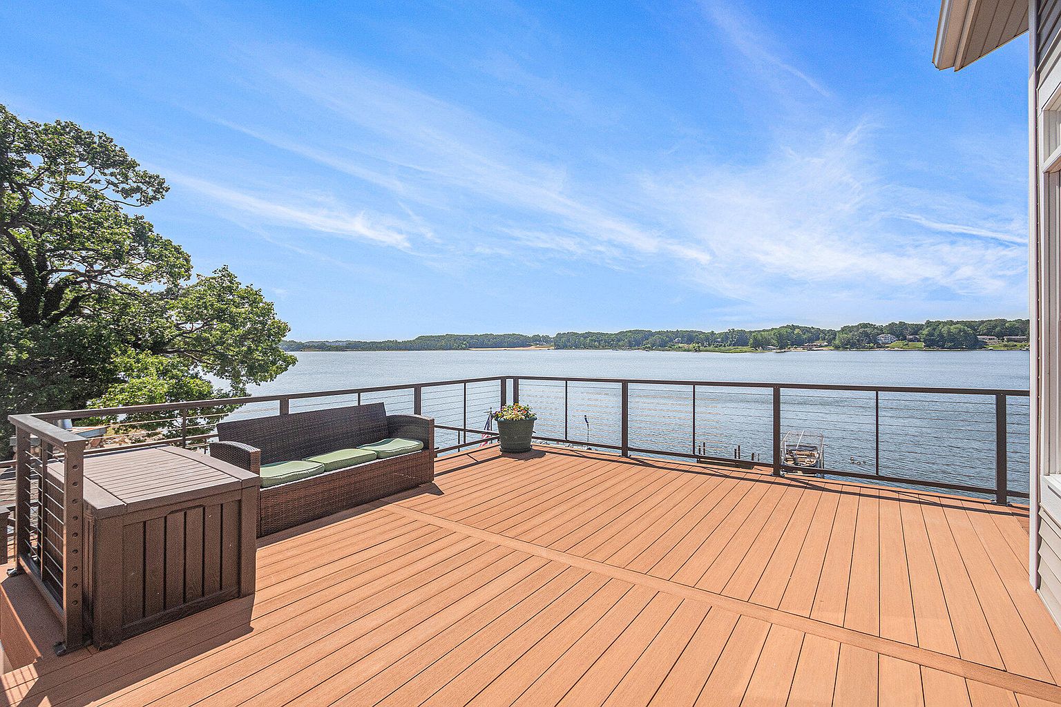 This image showcases a spacious wooden deck overlooking a serene lake. The deck features comfortable outdoor seating, including a wicker sofa with green cushions and a storage box, complemented by a modern railing system. The view extends to the opposite shore, creating a tranquil and inviting outdoor living space.