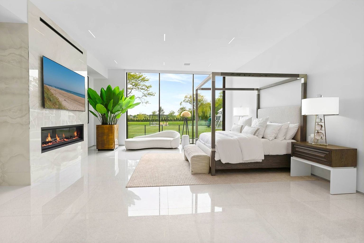 This expansive primary bedroom features a modern four-poster bed as the centerpiece, set against a backdrop of bright, neutral tones and polished light-colored flooring. A linear fireplace is integrated into a tall, stone-clad wall, while large windows provide an unobstructed view of the lush green backyard. The room is accented by a sculptural chaise lounge, a large potted plant, and a contemporary bedside table, creating a serene and luxurious atmosphere.