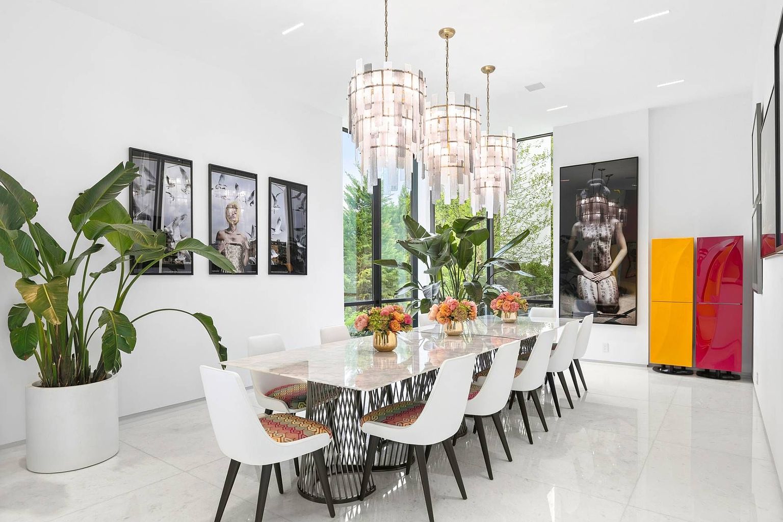 This bright and contemporary dining room features a long stone-topped table with a geometric metal base, surrounded by white chairs with colorful patterned cushions. Three tiered crystal chandeliers hang above the table, while large windows provide a view of the lush greenery outside. The space is accented by bold artwork on the walls and vibrant yellow and magenta storage cabinets, creating a sophisticated and artistic atmosphere.