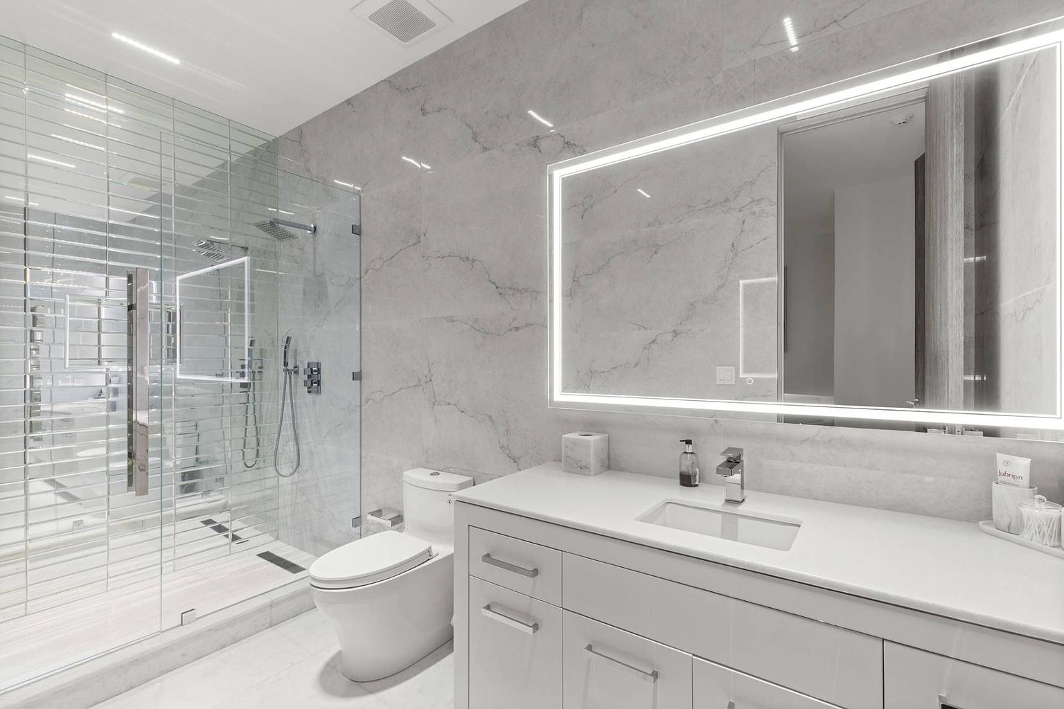 This modern primary bathroom features a sleek, minimalist aesthetic with large-format stone wall tiles and a clean white vanity. A glass-enclosed walk-in shower sits adjacent to a white toilet, while a large, illuminated rectangular mirror hangs above the sink. The space is bright and airy, characterized by its neutral color palette and contemporary fixtures.