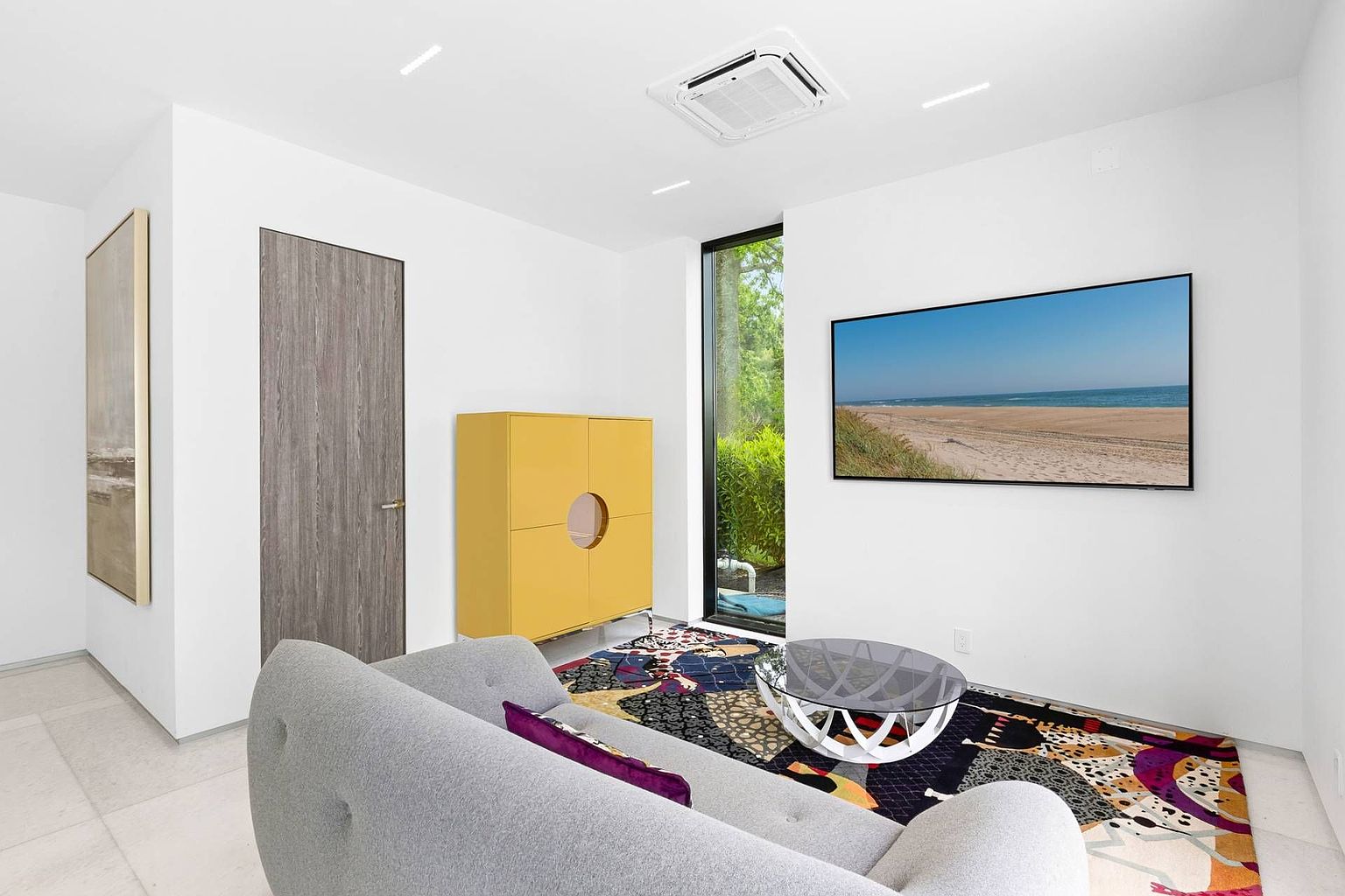 This modern living room features a minimalist aesthetic with crisp white walls and a light-colored tile floor. A grey sofa with a purple accent pillow faces a wall-mounted television displaying a beach scene, while a vibrant, multi-colored patterned rug anchors the seating area. A tall, yellow cabinet adds a bold pop of color next to a floor-to-ceiling window that provides a view of the lush greenery outside.