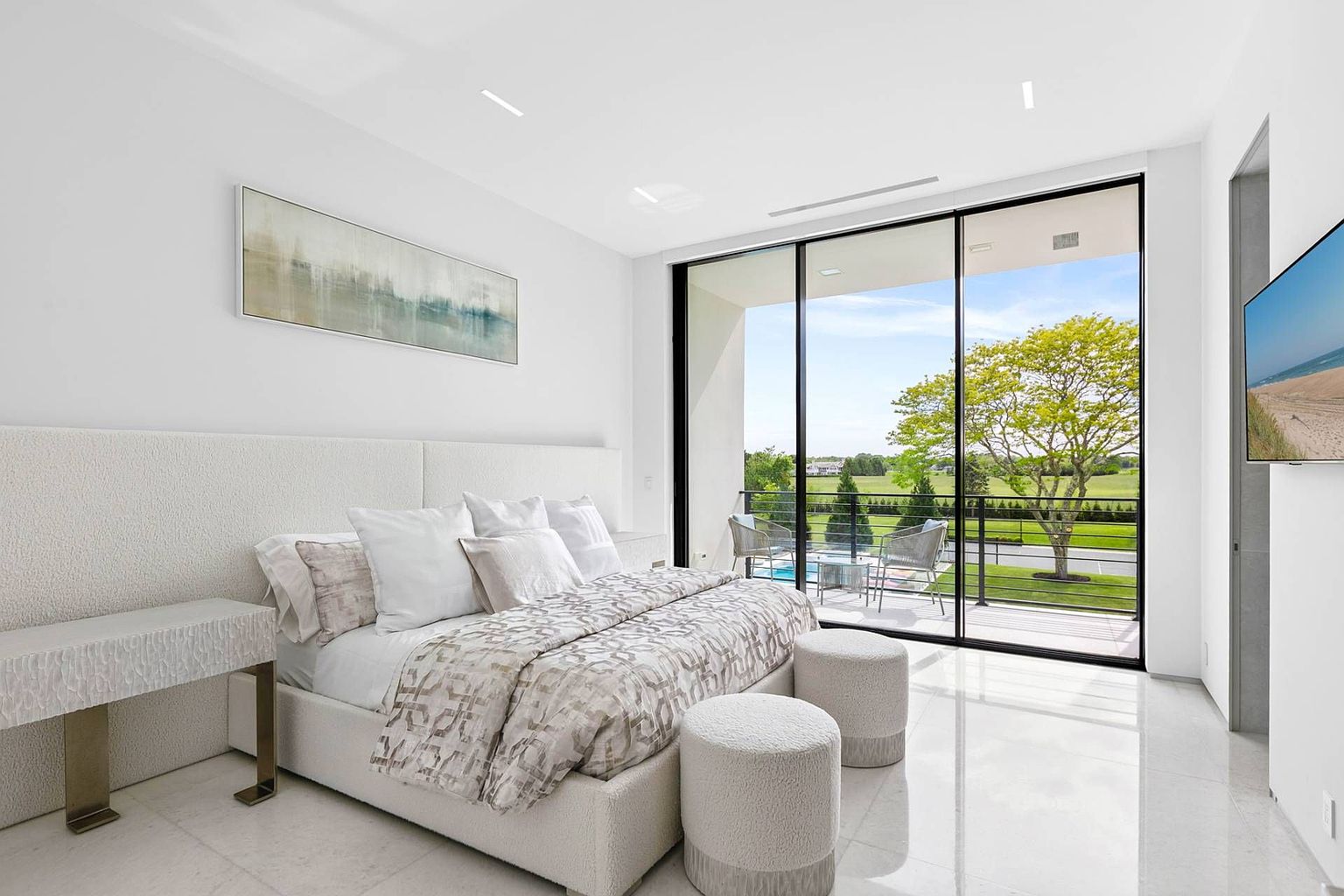 This bright and airy primary bedroom features a neutral color palette with a textured upholstered headboard and a patterned bedspread. Large floor-to-ceiling sliding glass doors open onto a private balcony, providing an expansive view of the lush green landscape and trees outside. The room is finished with polished light-colored flooring, two circular ottomans at the foot of the bed, and a sleek side table, creating a sophisticated and tranquil retreat.