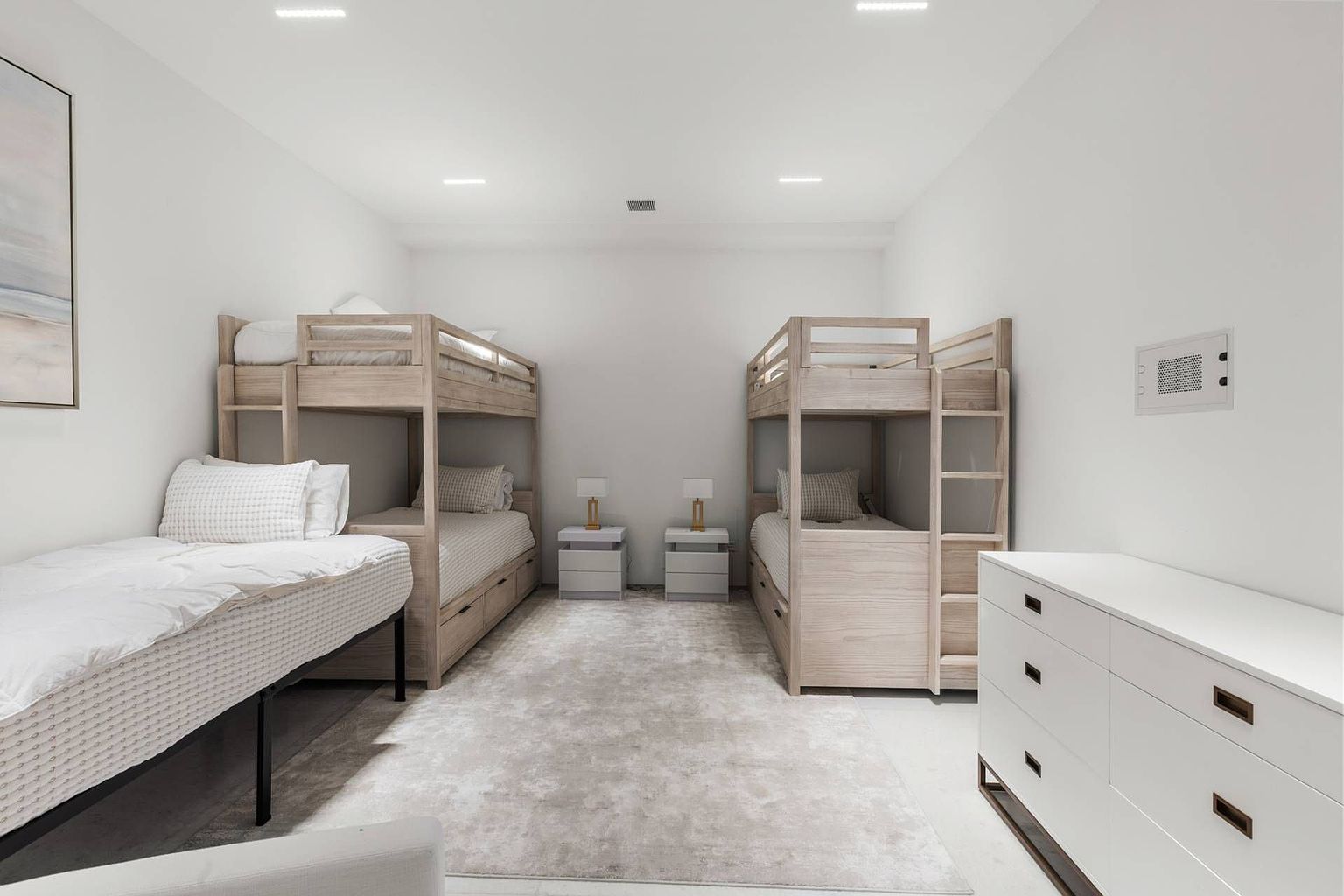 This spacious, minimalist bedroom is designed as a bunk room, featuring two light-wood bunk bed sets positioned on either side of a central area. A large, neutral-toned area rug anchors the space, while a white dresser provides storage on the right and a single bed sits in the foreground on the left. The room is brightly lit by recessed ceiling lights, creating a clean, airy, and functional atmosphere suitable for a guest or children's room.