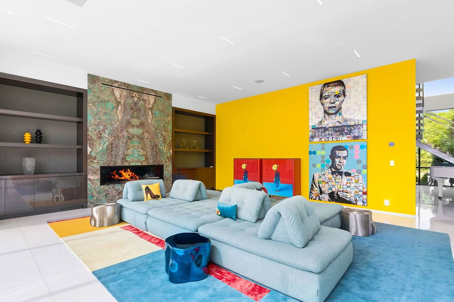 This vibrant living room features a large, light blue modular sectional sofa centered on a colorful area rug. A striking stone-clad chimney breast serves as a focal point, while the adjacent wall is painted a bold, bright yellow and adorned with two large pop-art portraits. The space is further accented by a glossy blue side table, a red cabinet, and built-in shelving, creating a modern and eclectic atmosphere.