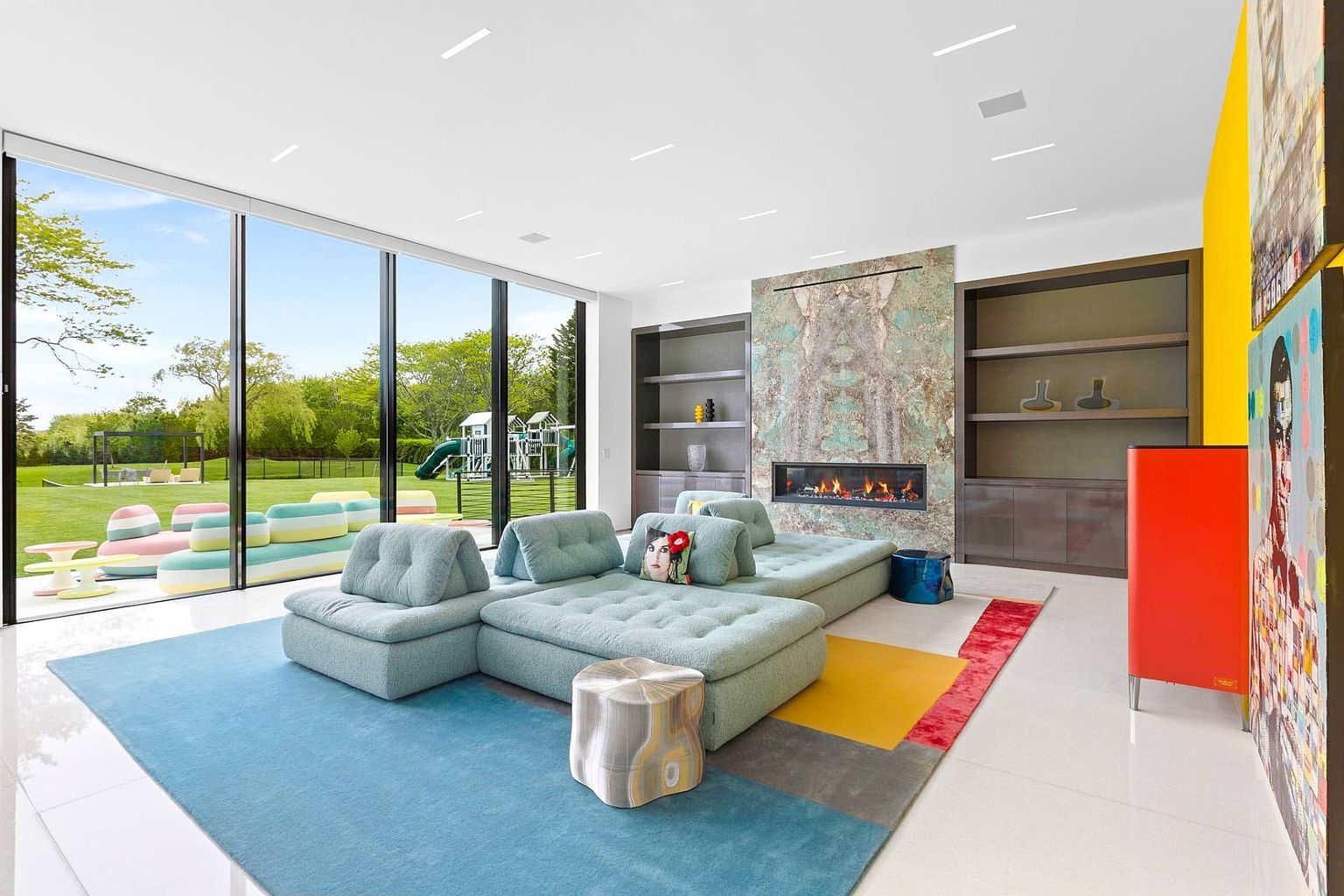 This contemporary living room features a large, light blue modular sofa set atop a vibrant, multi-colored area rug. A striking stone-clad chimney breast serves as a focal point, housing a wide linear fireplace between two built-in shelving units. The space is bathed in natural light from expansive floor-to-ceiling windows that provide a seamless view of the lush green backyard and playground.