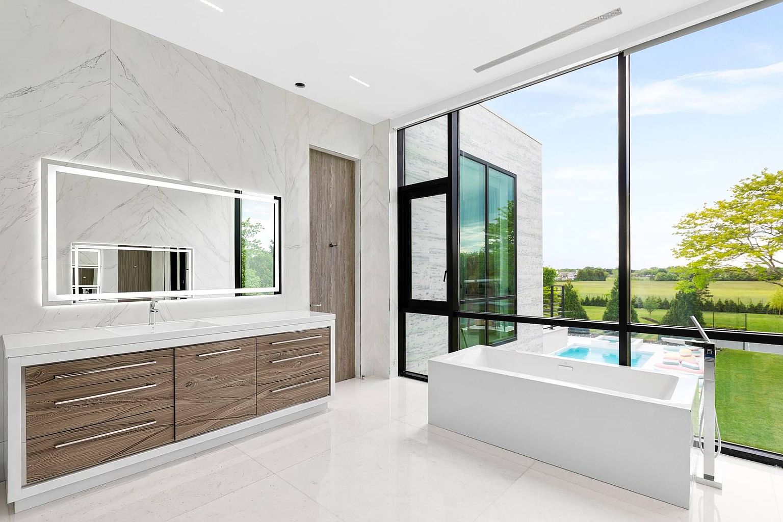 This luxurious primary bathroom features a sleek, modern aesthetic with a floating vanity, light wood-toned cabinetry, and a large, illuminated mirror. A freestanding rectangular soaking tub is positioned next to expansive, floor-to-ceiling windows that provide a panoramic view of the lush green landscape and a private pool area. The space is finished with light-colored, large-format tile flooring and walls, creating a bright, airy, and sophisticated atmosphere.