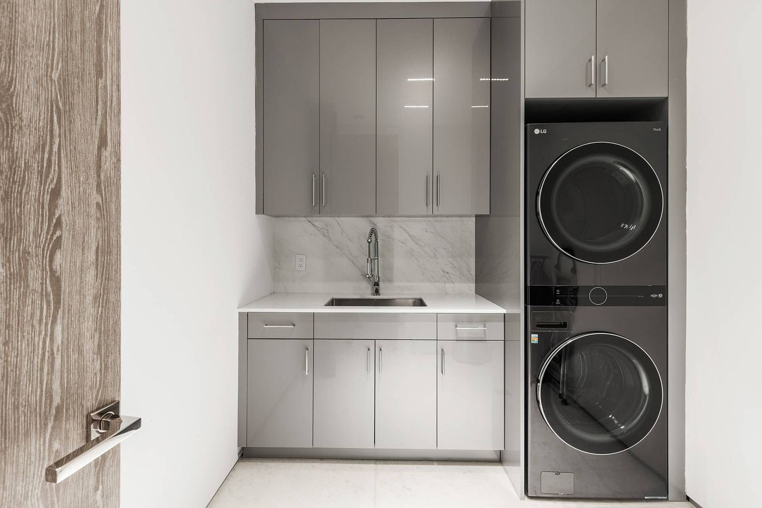 This modern laundry room features a sleek, stacked LG washer and dryer unit set within a custom cabinetry wall. The space includes a stone-topped counter with an integrated sink and a marble-patterned backsplash, complemented by glossy gray upper and lower cabinets. The clean, minimalist design is enhanced by bright lighting and neutral flooring, creating a functional and polished utility area.