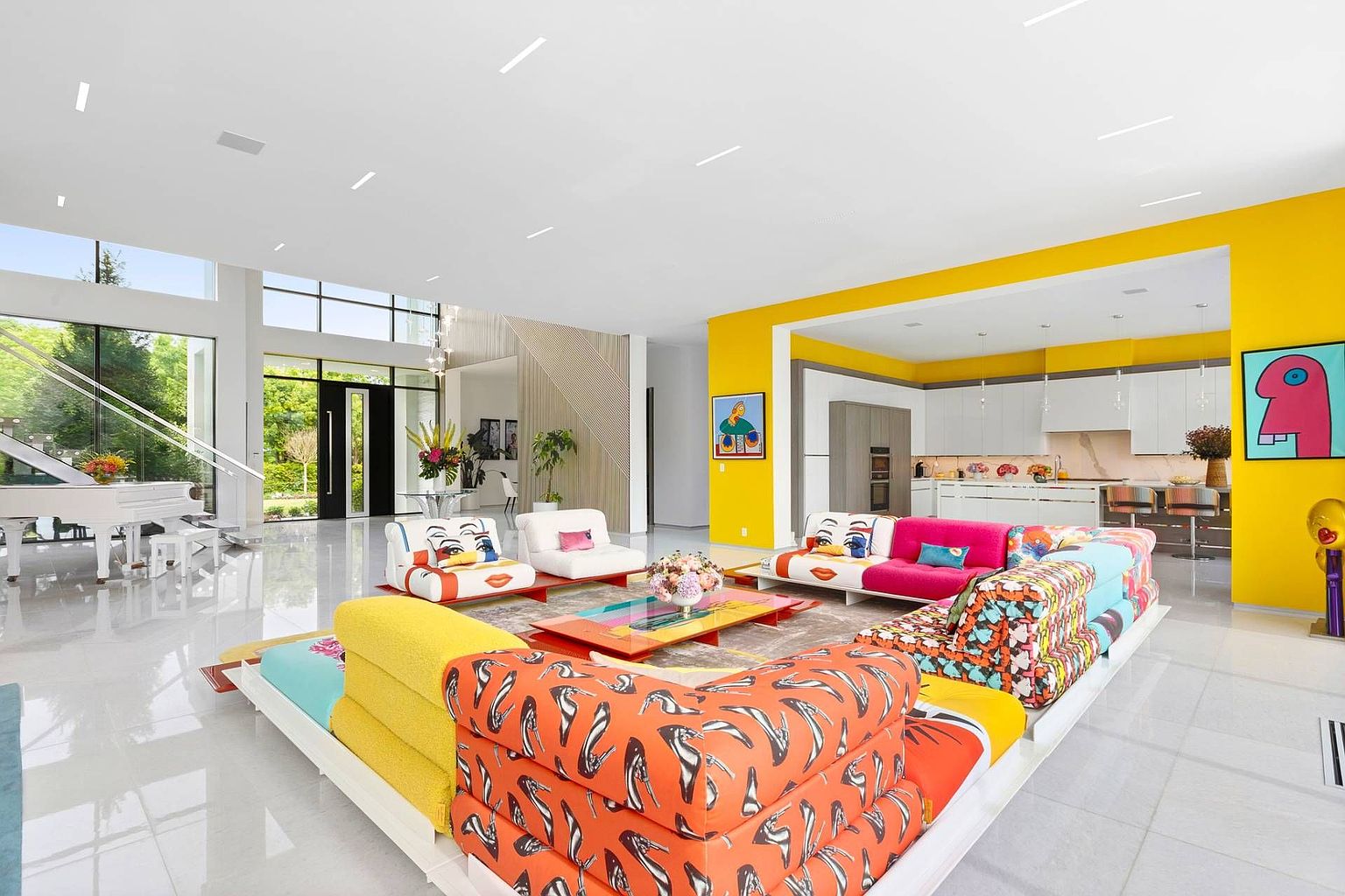 This expansive, open-concept living area features a vibrant, eclectic design with a large, multi-section modular sofa upholstered in bold, colorful patterns. The space is anchored by a glossy white floor and high ceilings, with a bright yellow accent wall framing the transition into a modern kitchen. A white grand piano sits near the tall windows, while contemporary art pieces add a playful, gallery-like aesthetic to the bright, airy room.