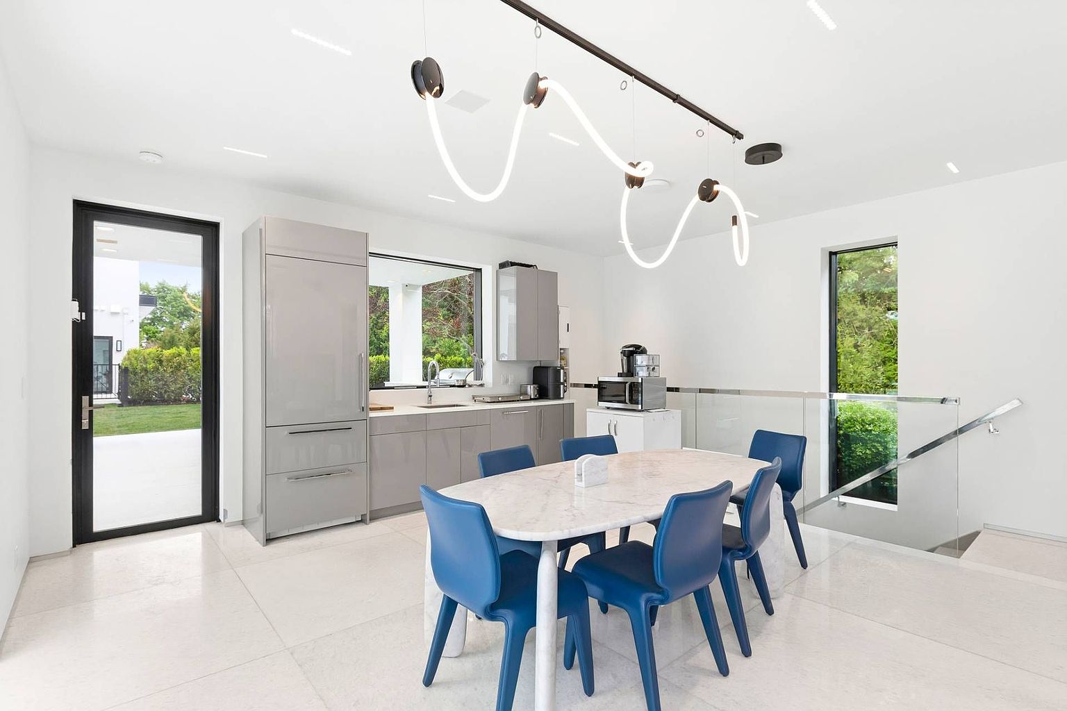 This bright, contemporary dining area features a sleek stone-topped table surrounded by six vibrant blue chairs. A unique, sculptural light fixture hangs above the table, serving as a focal point against the minimalist white walls and light-colored tile flooring. The space is open and airy, with a glass-railed staircase to the right and a functional kitchenette area with grey cabinetry and a window view to the left.