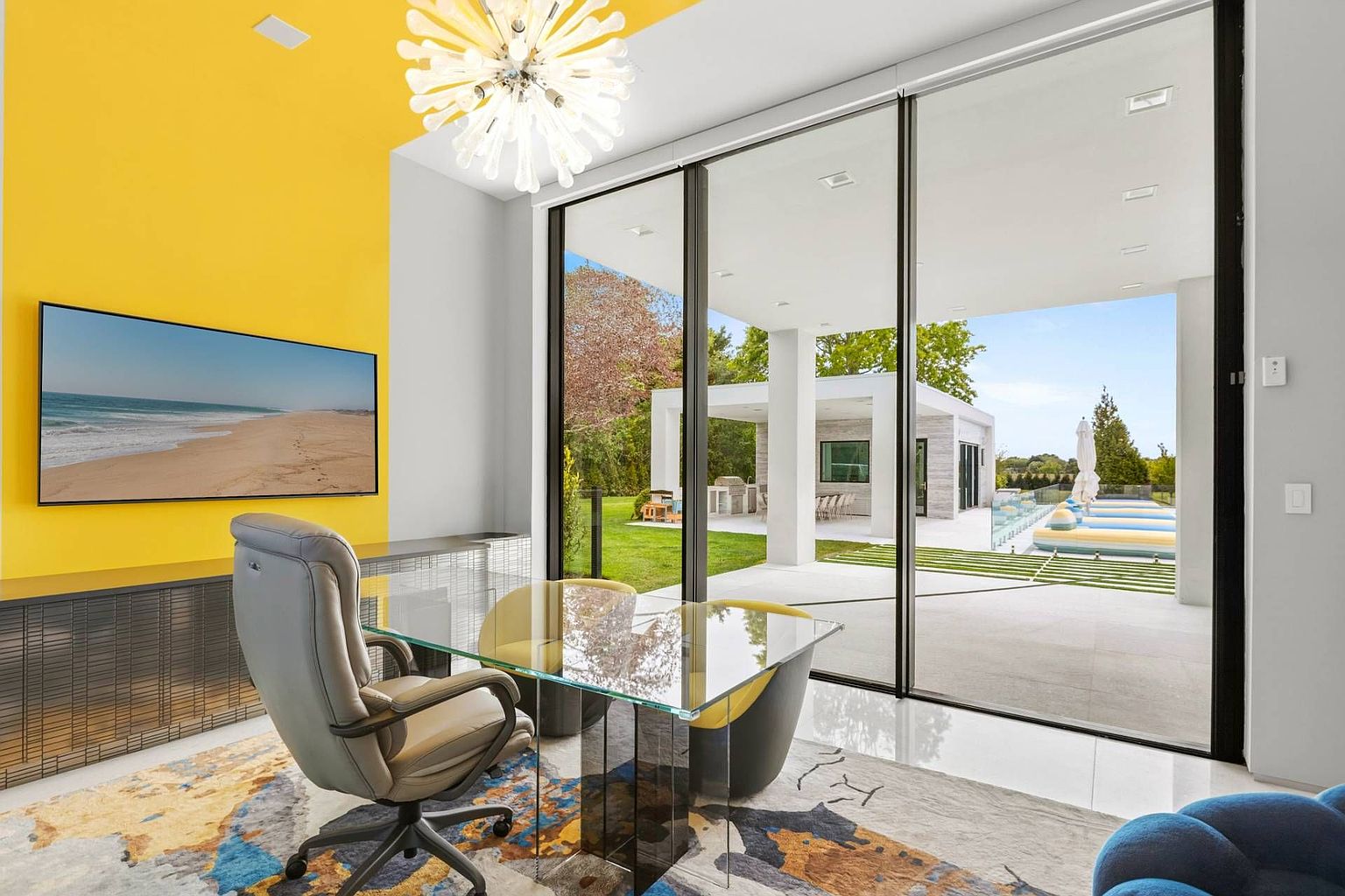 This modern home office features a bold yellow accent wall, a glass-topped desk, and a contemporary gray leather office chair. Large floor-to-ceiling glass sliding doors provide a seamless transition to an outdoor patio and pool area, bathing the room in natural light. The space is anchored by a vibrant, abstract-patterned area rug and illuminated by a unique, starburst-style chandelier.