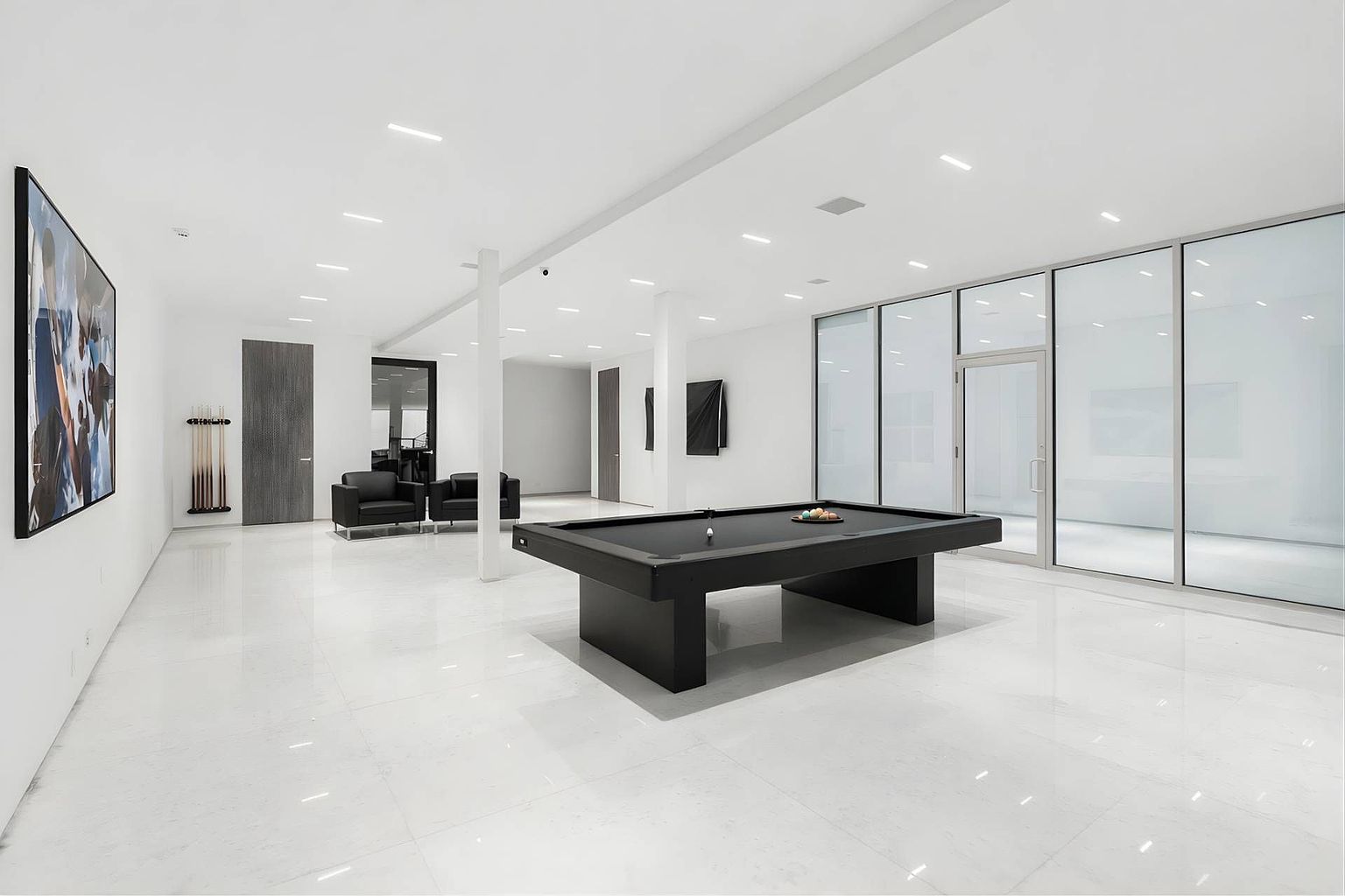 This minimalist game room features a sleek black pool table as the central focal point, set against a backdrop of stark white walls and polished light-colored flooring. To the left, a pair of black leather armchairs and a wall-mounted cue rack provide a sophisticated lounge area, while a wall of floor-to-ceiling glass panels separates the space from an adjacent room. The room is illuminated by recessed lighting, creating a clean, modern, and gallery-like atmosphere.