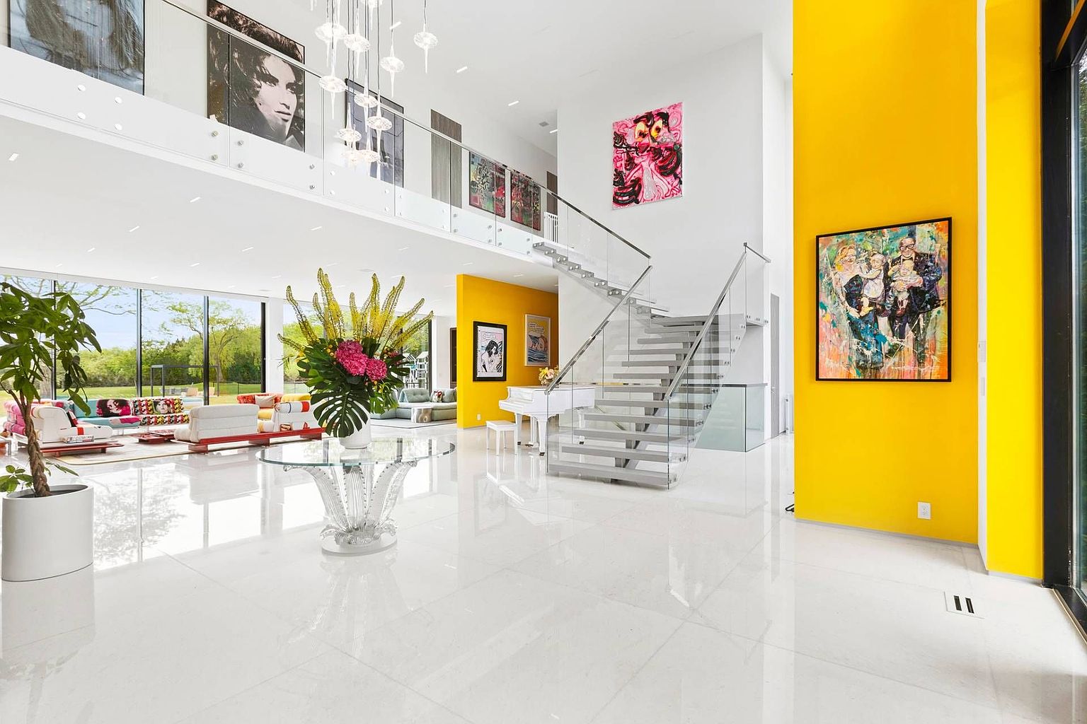 This expansive, double-height living area features a striking combination of white surfaces and bold yellow accent walls, creating a vibrant and modern aesthetic. A glass-railed staircase serves as a central architectural feature, while large windows provide an abundance of natural light and views of the outdoor landscape. The space is decorated with contemporary art and a grand piano, emphasizing a luxurious and open-concept design.