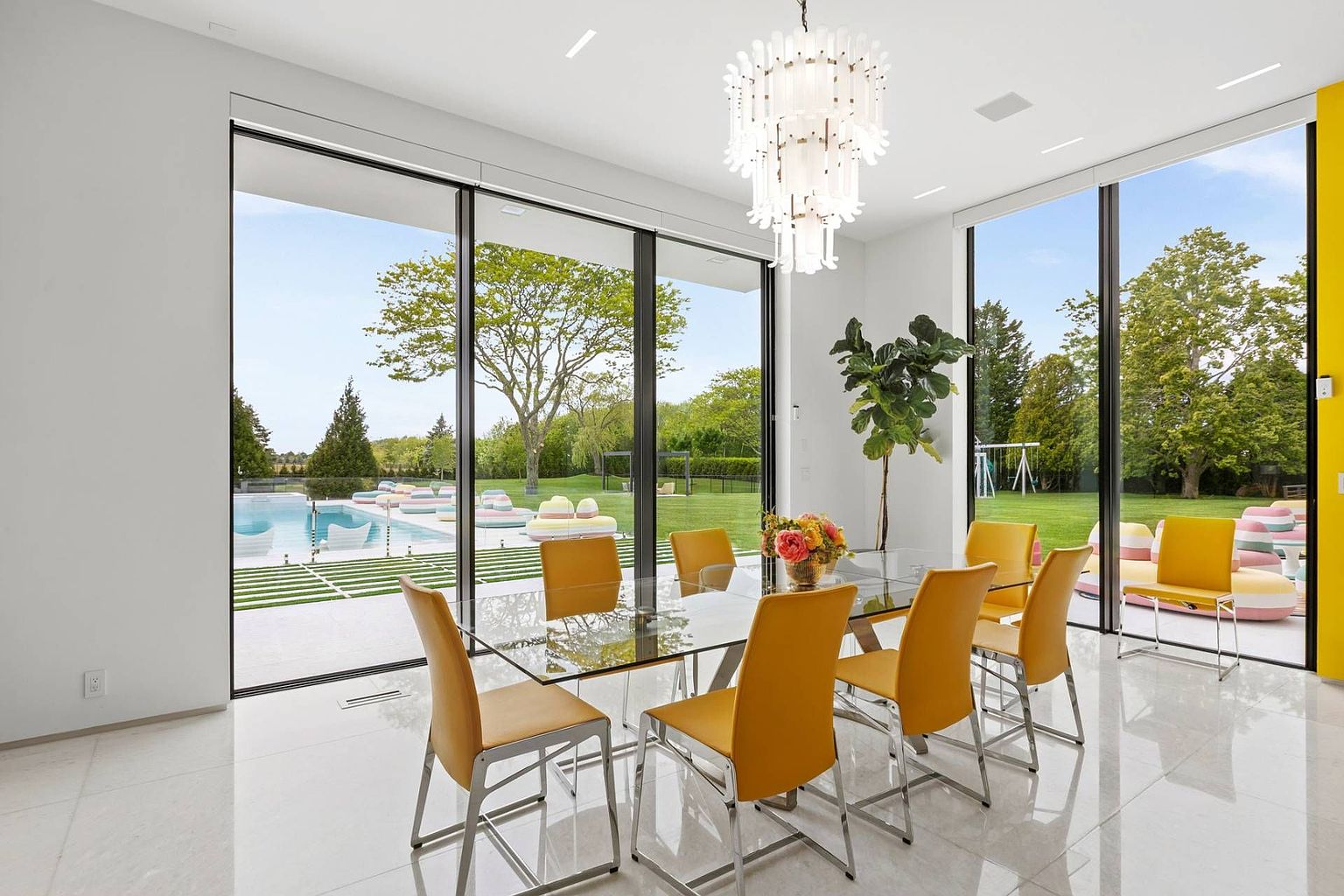 This bright and airy dining room features a glass-topped table surrounded by vibrant yellow chairs, set beneath a striking, multi-tiered glass chandelier. Large floor-to-ceiling windows provide an expansive view of the lush backyard and swimming pool, creating a seamless connection between the indoor and outdoor spaces. The room is finished with polished light-colored flooring and minimalist white walls, emphasizing a clean, contemporary aesthetic.