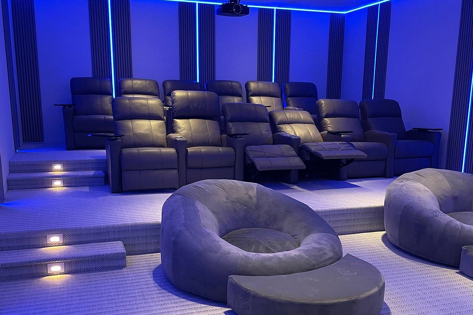 This image showcases a dedicated home theater room featuring tiered seating with multiple rows of dark leather recliners. The foreground includes two plush, circular lounge chairs with matching ottomans, while the walls are accented with vertical slats and integrated blue LED strip lighting. The room is designed for an immersive cinematic experience, with a projector mounted on the ceiling and recessed step lighting along the tiered platforms.