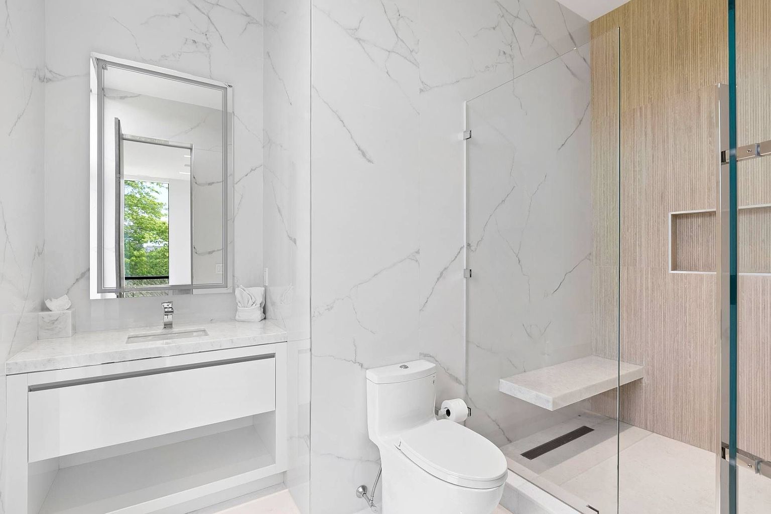This modern bathroom features a sleek vanity with a stone countertop and a large rectangular mirror reflecting a bright window. To the right, a glass-enclosed shower area includes a built-in stone bench and a linear drain, set against a backdrop of light-toned wood-look wall paneling. The space is finished with large-format stone wall tiles, creating a clean, minimalist, and high-end aesthetic.