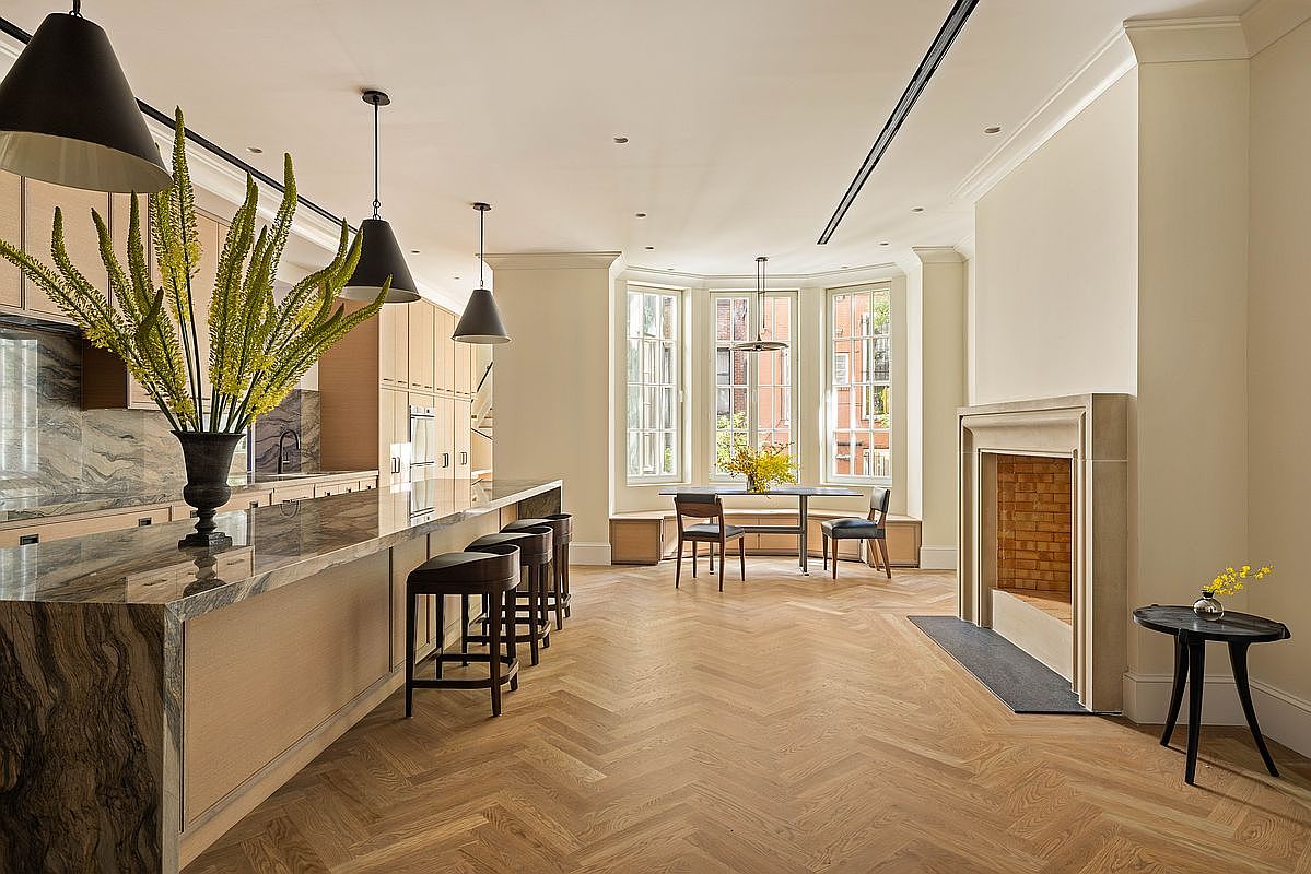 This spacious, open-concept kitchen features a large island with a stone countertop and integrated seating, complemented by light wood cabinetry and a herringbone-patterned wood floor. A stone-clad fireplace adds a focal point to the room, while a dining area is positioned near a set of tall windows that bring in natural light. The perspective is a wide-angle shot from the kitchen island, showcasing the seamless flow between the cooking, dining, and living areas.