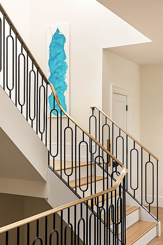 This interior view captures a staircase featuring a distinctive black metal railing with an elongated oval pattern, topped by a warm-toned wood handrail. The light-colored walls provide a neutral backdrop for a vibrant, textured blue abstract art piece mounted vertically on the wall. The perspective looks up the staircase, highlighting the clean lines and modern architectural design of the home's circulation space.