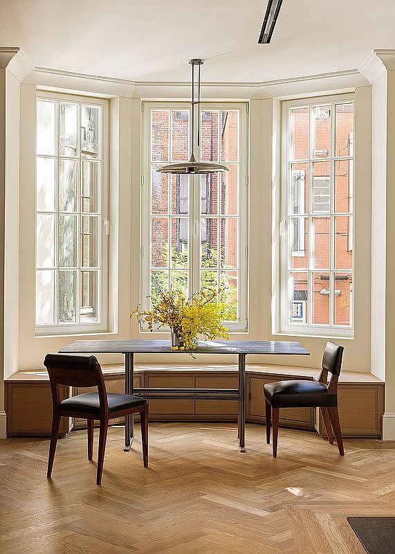 This elegant dining area features a bay window configuration with tall, multi-pane windows that flood the space with natural light. A sleek, dark-toned table is centered in the room, flanked by two modern chairs with dark leather upholstery, all set atop a light-toned herringbone wood floor. Built-in cabinetry runs along the base of the windows, providing a seamless and functional storage solution beneath the bright, airy breakfast nook.