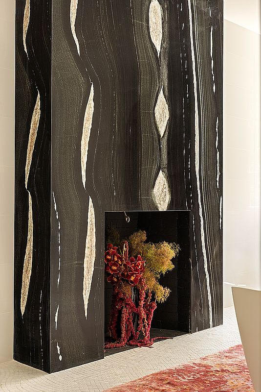 This image highlights a striking, tall fireplace surround crafted from dark, heavily veined stone with dramatic, light-colored vertical streaks. The fireplace opening is filled with a vibrant, textured floral arrangement in deep red and golden tones, creating a bold focal point. The surrounding area features a light-colored, textured floor, adding a subtle contrast to the dark, imposing stone structure.