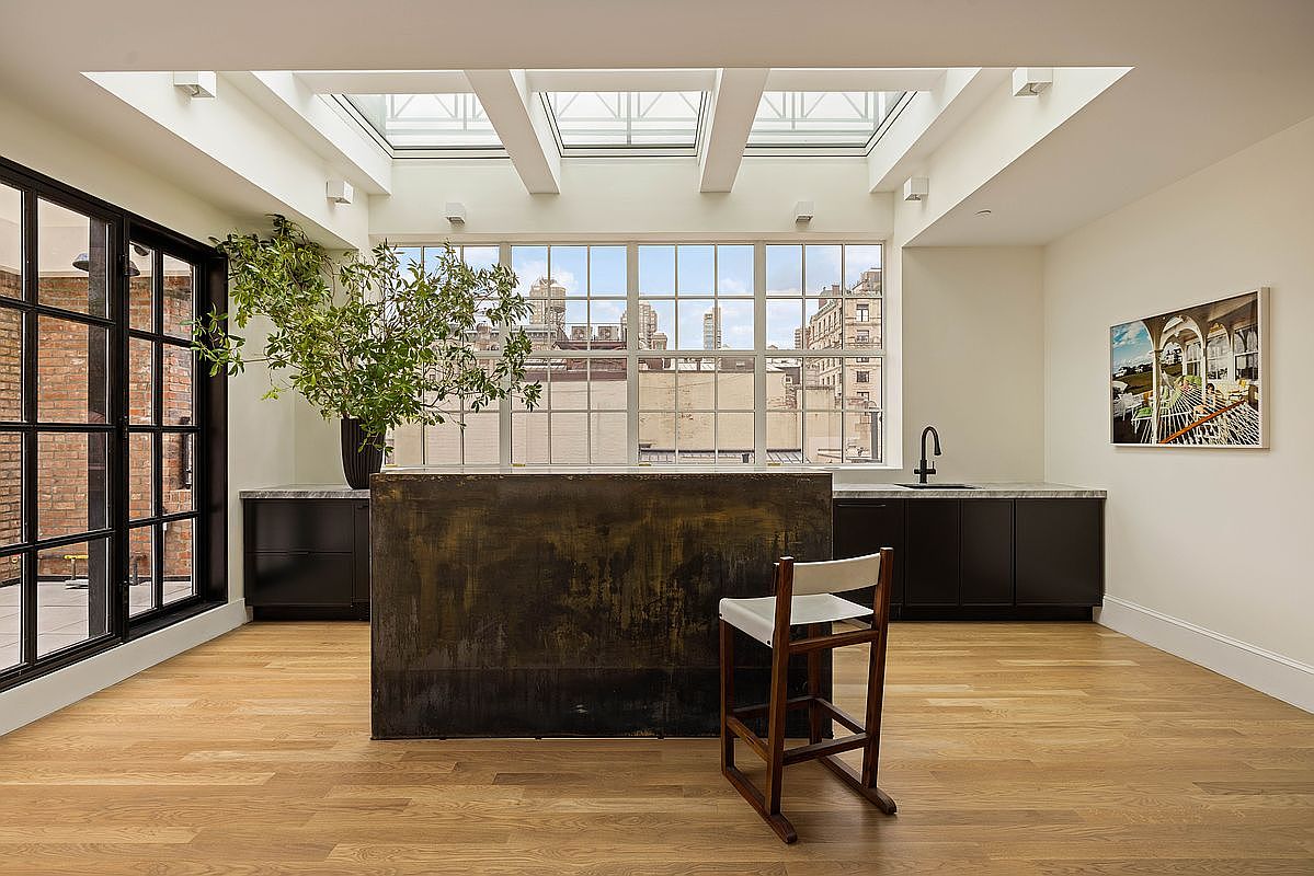 This sophisticated kitchen space features a prominent, dark-toned metal island as its centerpiece, complemented by a single wooden chair. The room is bathed in natural light from a series of skylights and a large, multi-pane window that overlooks an urban cityscape. Dark cabinetry with stone countertops lines the back wall, creating a sleek, modern aesthetic against the warm light wood flooring.