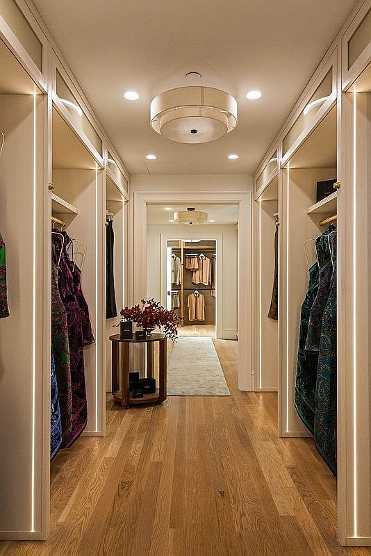 This luxurious walk-in closet features custom light wood cabinetry with integrated vertical LED lighting strips that illuminate the hanging garments. A central round table with a floral arrangement sits atop a light-toned runner, leading the eye through a doorway into a secondary, equally organized closet space. The warm wood flooring and recessed ceiling lighting create a sophisticated and inviting atmosphere.