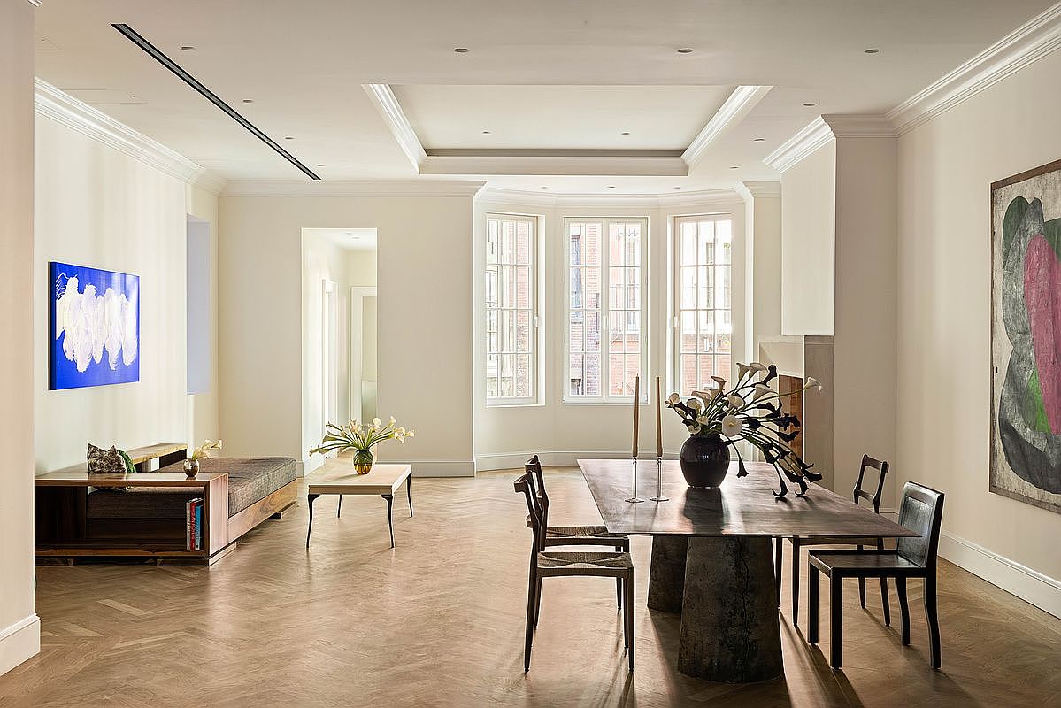 This elegant, open-concept space features a dining area in the foreground with a dark, sculptural table and matching chairs, complemented by a nearby living area with a low-profile sofa. The room is characterized by light-toned walls, herringbone-patterned wood flooring, and a bay window arrangement that fills the space with natural light. Large, bold abstract art pieces on the walls add a contemporary touch to the sophisticated, minimalist interior design.