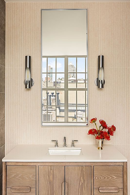 This elegant bathroom vanity area features a light wood cabinet with integrated handles, topped by a clean white counter and a centered sink. A tall, rectangular mirror is flanked by two modern, cylindrical wall sconces, reflecting a view of city buildings through the window. A small vase with vibrant red flowers adds a pop of color to the neutral, textured wall backdrop.
