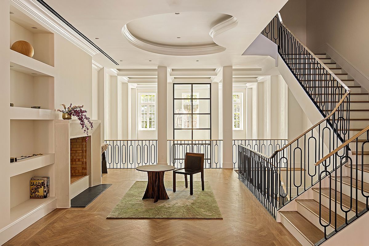 This elegant landing area features a sophisticated open-plan design with a grand staircase featuring a dark metal railing and a light wood handrail. The space is anchored by a circular table and a single chair set upon a green area rug, positioned before a large, grid-paned window that overlooks the space below. Architectural details include a recessed ceiling cove, built-in shelving units, and a fireplace with a clean-lined surround, all finished in a neutral palette that emphasizes the herringbone wood flooring.