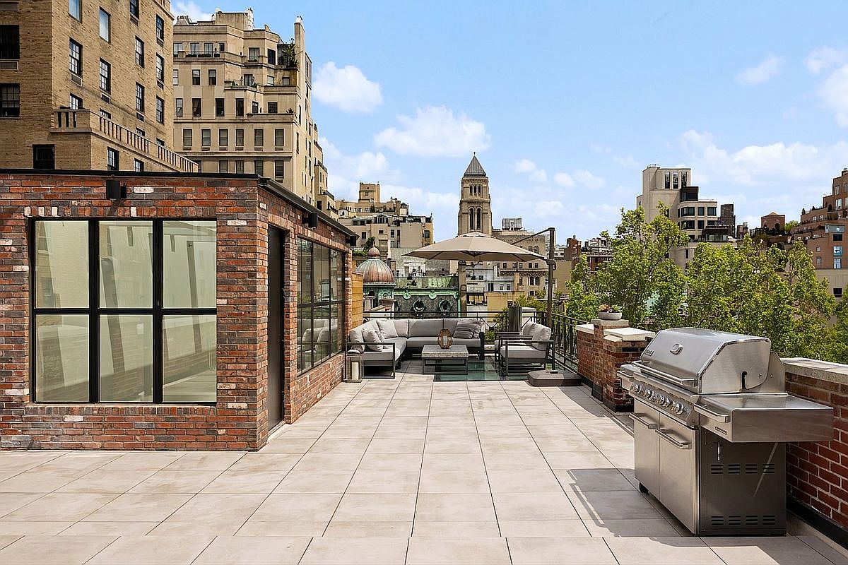 This expansive rooftop terrace offers a sophisticated outdoor living space set against a backdrop of urban architecture. The area features a modern sectional seating arrangement shaded by a large umbrella, complemented by a stainless steel grill station on the right. The space is finished with light-toned pavers and framed by a brick structure with large, multi-paned windows, creating a seamless blend of industrial charm and contemporary comfort.