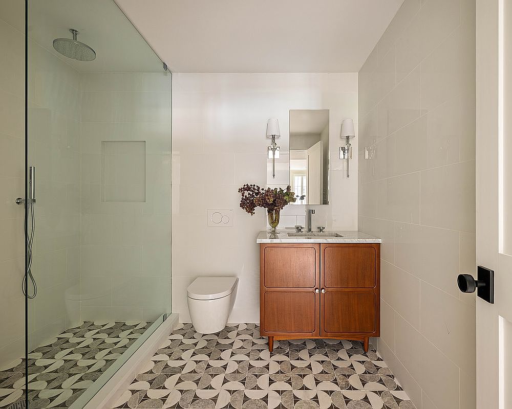This modern bathroom features a striking geometric patterned tile floor that contrasts with the clean, white tiled walls. A mid-century inspired wood vanity with a stone countertop sits beneath a rectangular mirror flanked by two wall sconces. To the left, a glass-enclosed walk-in shower with a rainfall showerhead completes the compact and stylish layout.