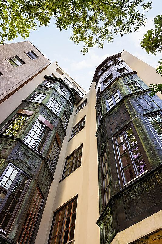 This low-angle, upward-looking perspective captures the striking architectural details of a multi-story building's rear facade. The structure features prominent, vertically oriented bay windows clad in weathered, oxidized metal panels that contrast against the smooth, light-colored stucco walls. The composition emphasizes the geometric complexity of the building's design, framed by the soft green canopy of overhanging tree branches against a bright sky.