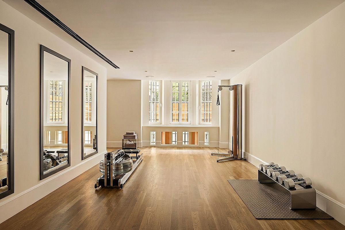This bright and spacious home gym features light wood flooring and neutral walls, creating a clean and inviting workout environment. The room is equipped with a rowing machine, a cable pulley system, and a rack of dumbbells on a rubber mat, while large mirrors on the left wall enhance the sense of space. Three sets of tall windows at the far end of the room allow for ample natural light, providing a serene view of the outdoors.