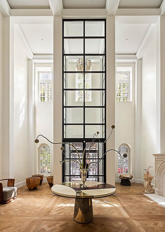 This grand, double-height entryway features a striking central grid-patterned window wall that serves as a dramatic focal point. The space is anchored by a sculptural, multi-material pedestal table topped with an artistic floral arrangement, set against a backdrop of light-toned walls and intricate herringbone wood flooring. Arched windows on either side and minimalist wooden stools contribute to an atmosphere of sophisticated, architectural elegance.