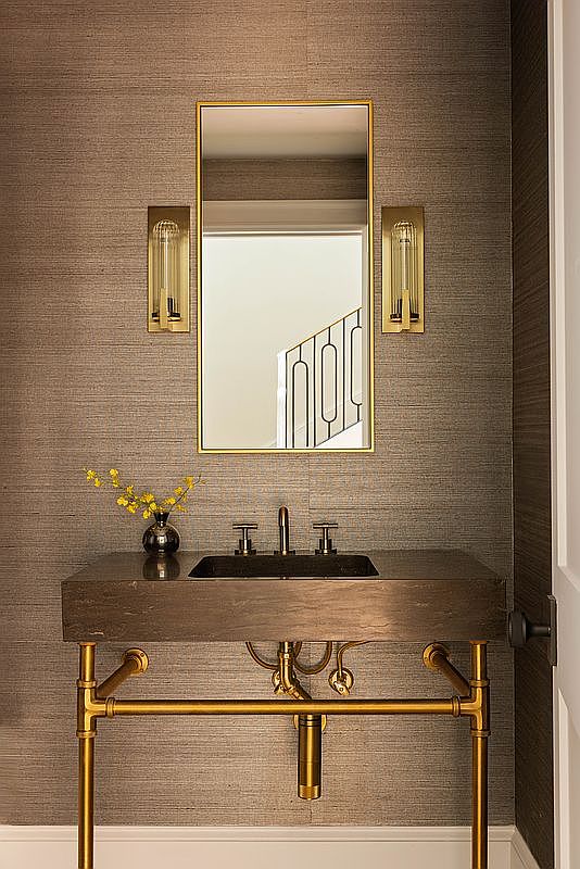 This sophisticated powder room features a wall-mounted stone vanity with a dark, textured basin supported by a polished brass pipe-style frame. The walls are covered in a rich, textured wallpaper, complemented by a rectangular mirror flanked by two elegant, vertical brass wall sconces. A small vase with yellow flowers adds a touch of color to the vanity, creating a refined and modern aesthetic.