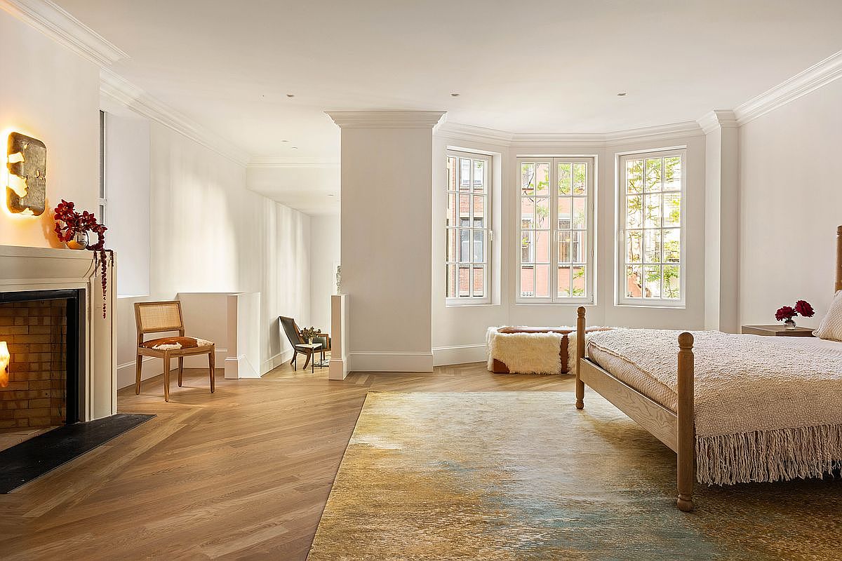 This spacious primary bedroom features light wood herringbone flooring and a neutral color palette that creates a serene atmosphere. A prominent fireplace with a classic mantel serves as a focal point on the left, while a large bed with a textured throw sits on the right atop a vibrant, abstract-patterned area rug. The room is bathed in natural light from a set of tall windows, and a small seating area with a cane-back chair adds a touch of refined elegance to the corner.