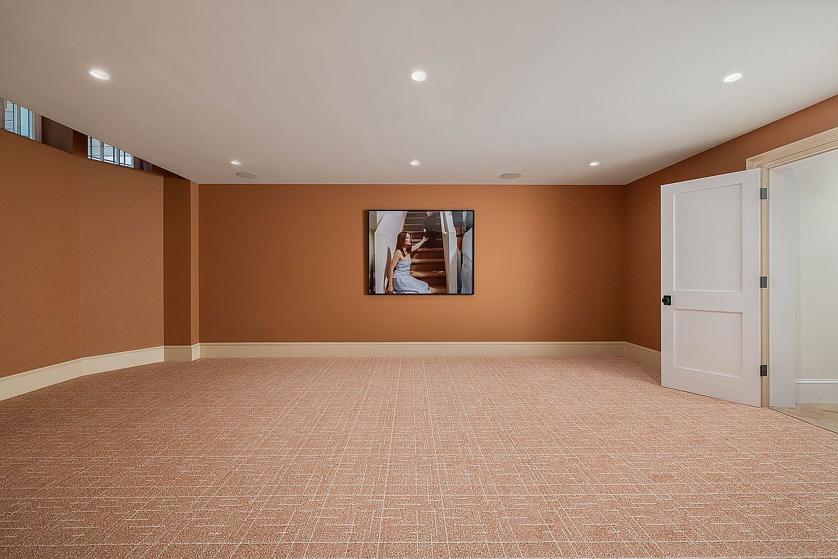 This spacious, minimalist basement room features warm, terracotta-toned walls and a textured, light-colored patterned carpet. A single piece of framed artwork hangs centered on the main wall, while recessed ceiling lighting provides even illumination throughout the space. A white door on the right leads to an adjacent area, and small, high-set windows are visible on the upper left wall.
