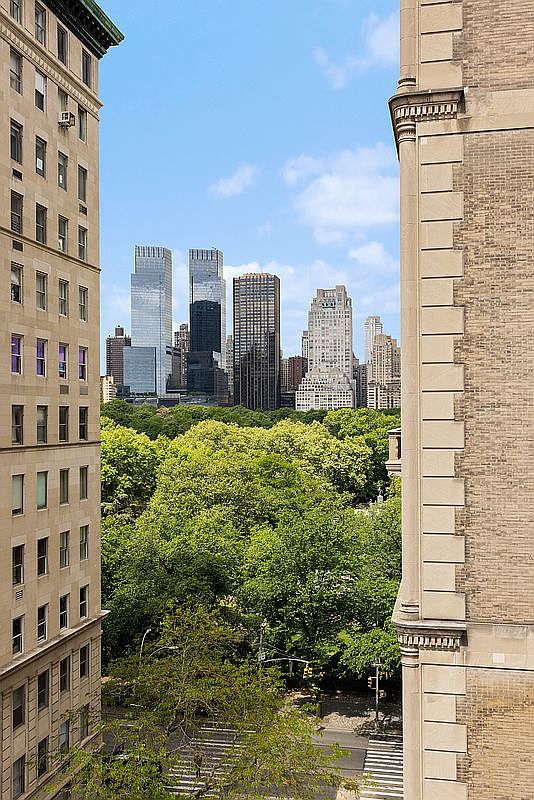 This elevated perspective captures a classic urban view framed by two historic stone-clad buildings, looking out over a lush, dense canopy of trees in Central Park. In the distance, a collection of iconic New York City skyscrapers rises against a bright blue sky with scattered clouds. The composition emphasizes the contrast between the architectural masonry of the foreground buildings and the vibrant greenery of the park below.