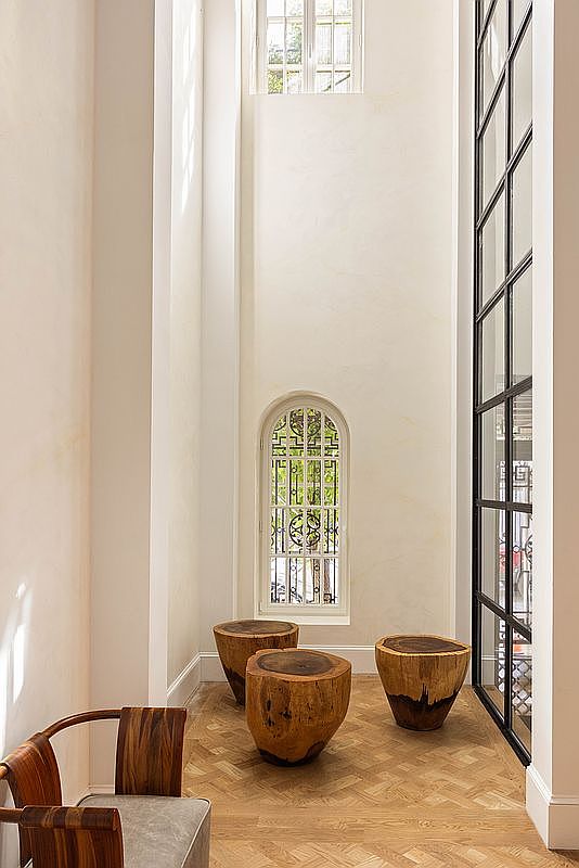 This elegant, double-height entryway features light-toned walls and a classic herringbone-patterned wood floor. The space is accented by three organic, raw-edge wood stump tables and a modern chair, with a tall, arched window featuring decorative ironwork. A large, black-framed glass partition runs vertically along the side, adding a contemporary architectural element to the bright, airy foyer.