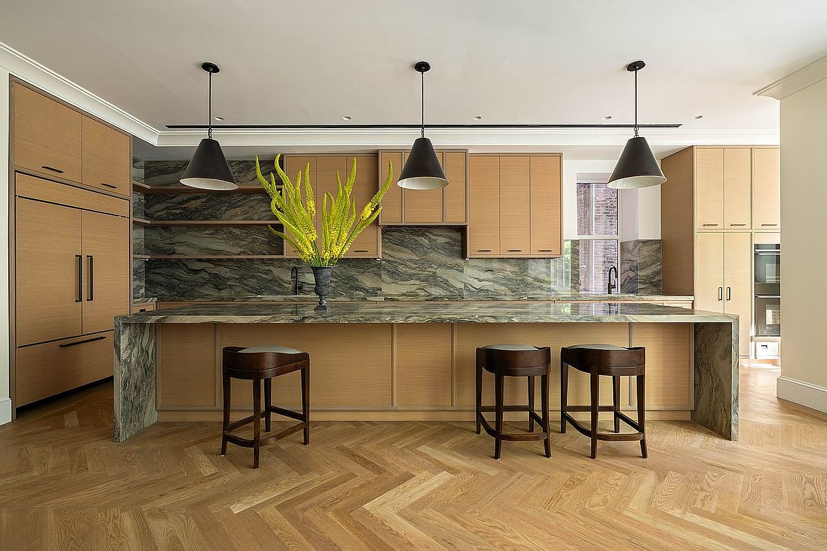 This sophisticated kitchen features a large central island with a waterfall-edge stone countertop and three dark wood stools. The space is defined by light wood cabinetry, a striking stone backsplash, and a herringbone-patterned wood floor. Three pendant lights hang above the island, creating a warm and inviting atmosphere in this modern, high-end culinary space.