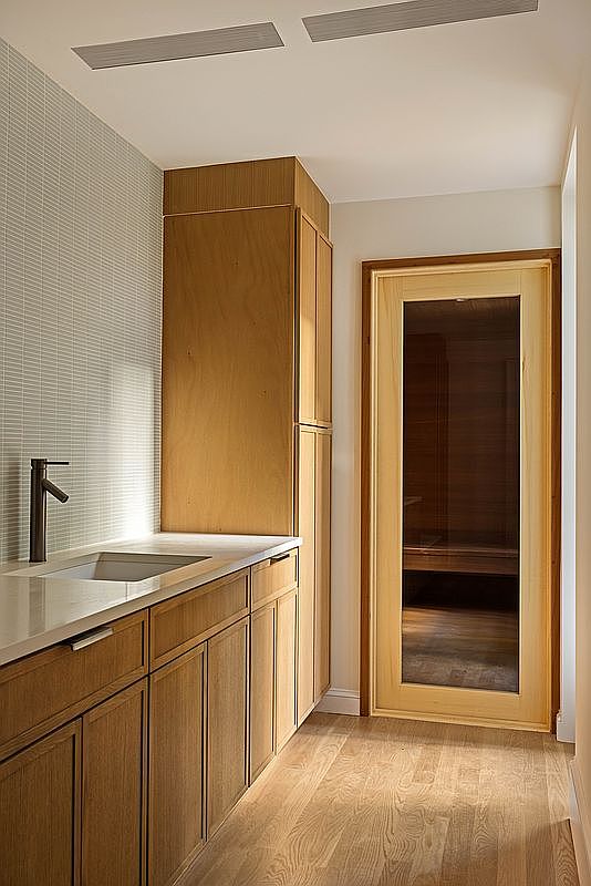 This interior space features a sleek vanity with a light wood finish, a stone countertop, and a modern faucet, set against a wall with textured, light-colored tile. To the right, a glass-paneled door leads into a sauna, creating a spa-like atmosphere. The room is finished with light wood flooring and recessed ceiling lighting, offering a clean and contemporary aesthetic.
