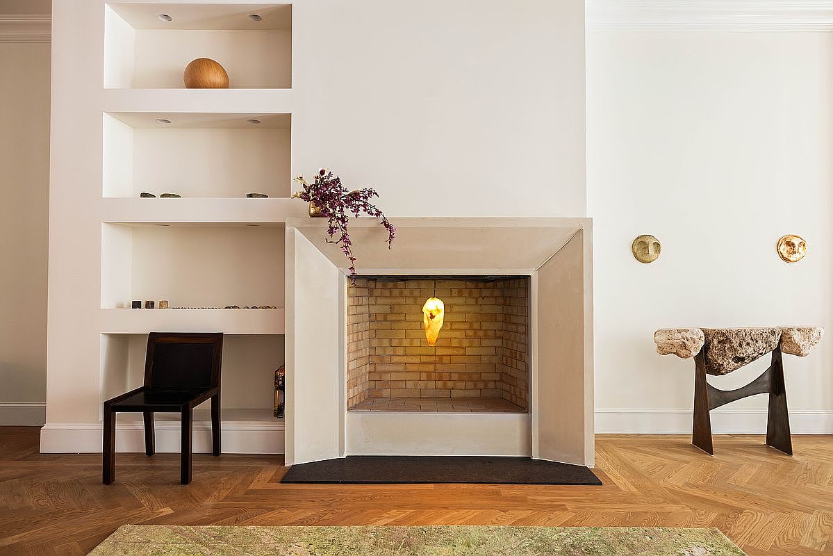 This sophisticated living room features a prominent fireplace with a minimalist, light-toned surround and a brick-lined firebox containing a unique hanging light fixture. To the left, built-in shelving provides display space, while a dark wood chair sits nearby on the herringbone-patterned wood flooring. To the right, a sculptural console table made of raw stone and dark metal adds an artistic touch, complemented by two circular wall accents.