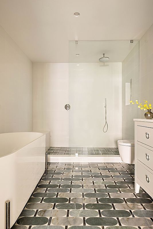 This modern bathroom features a sleek freestanding soaking tub and a spacious walk-in shower enclosed by a glass panel. The floor is covered in a distinctive patterned tile with rounded rectangular shapes, creating a sophisticated visual texture. A white vanity with silver hardware and a small vase of yellow flowers adds a bright, clean touch to the space.