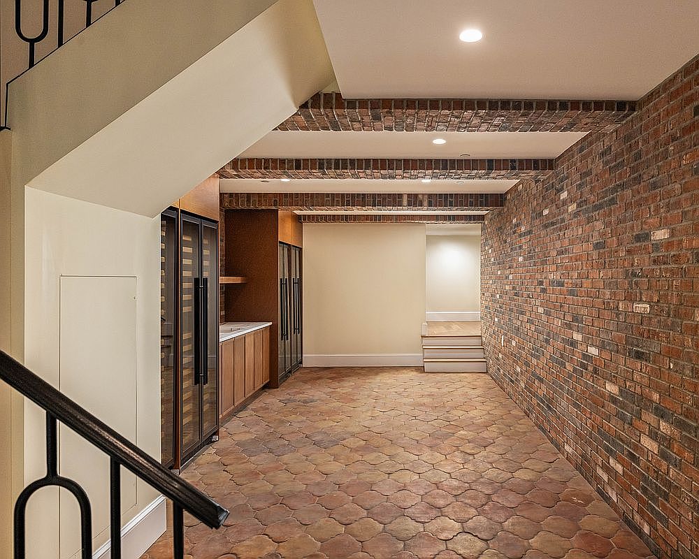 This sophisticated wine cellar features a distinctive terracotta-toned hexagonal tile floor and a prominent exposed brick wall that runs the length of the space. The ceiling is accented with decorative brick arches, while a sleek, modern wet bar area with glass-front wine refrigeration units is tucked neatly under the staircase. The perspective captures the transition from the foreground staircase down into the cellar, highlighting a warm, rustic-industrial aesthetic.
