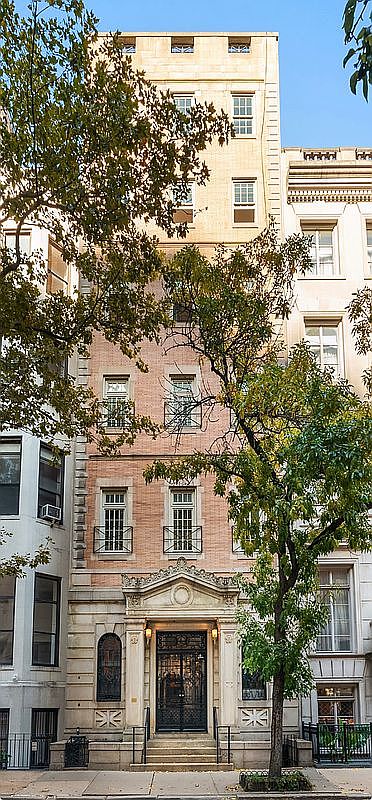 This image captures the front facade of a multi-story townhouse, showcasing a classic architectural style with a brick exterior and a prominent, ornate stone entryway. The building features symmetrical window placement, decorative iron railings on the lower levels, and a distinct transition to a lighter-colored stone facade on the upper floors. A mature tree stands in the foreground, partially framing the structure and adding a sense of scale to the urban streetscape.