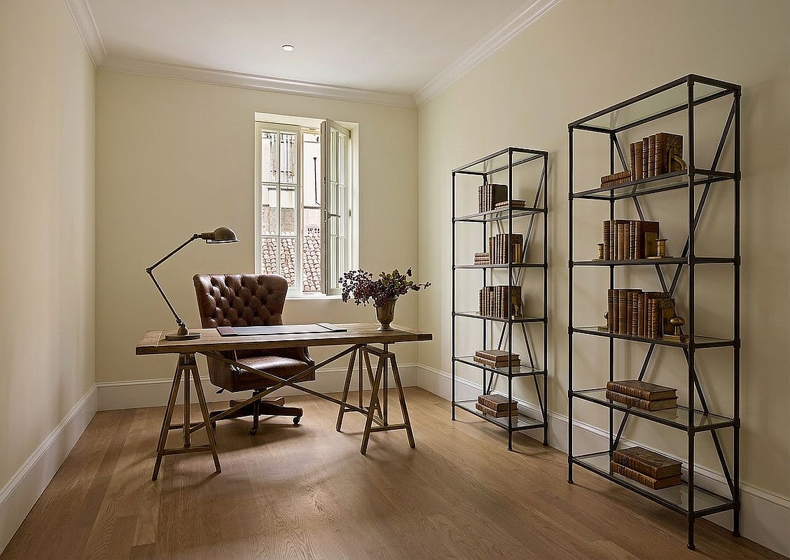 This sophisticated home office features a classic leather tufted chair paired with a rustic wood desk on a metal trestle base. Two tall, industrial-style metal shelving units with glass tiers provide ample storage for books, while a window allows natural light to brighten the neutral-toned room. The space is finished with light wood flooring and clean, minimalist walls, creating a professional and serene atmosphere.
